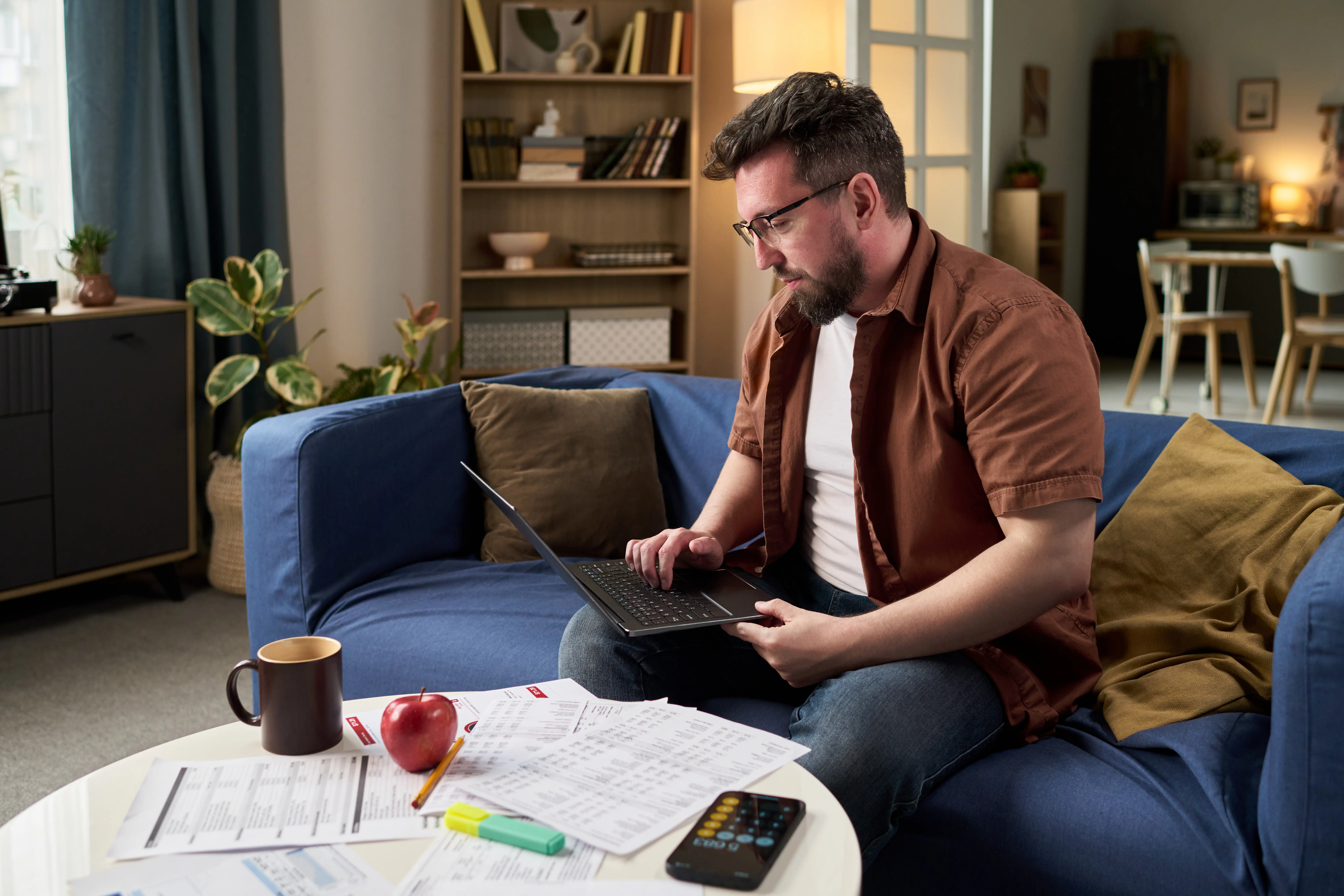 Ein junger erwachsener kaukasischer Mann berechnet Steuern mit einem Laptop und Papierdokumenten.